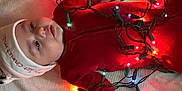 Oaklynn joined the competition — help win amazing prizes! baby, child, christmas_lights, red_onesie, white_hat, blanket, indoor, holiday, festive, toddler, cute, face, smiling, lying_down, cozy, celebration, colorful_lights, person, infant, warm