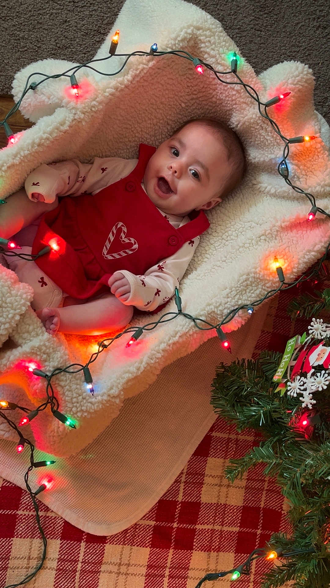 Oaklynn joined the competition — help win amazing prizes! baby, infant, blanket, christmas_lights, holiday, red_clothing, candy_cane, face, smiling, happy, festive, decorations, plaid, carpet, greenery, christmas_tree, indoors, cozy, cute, person