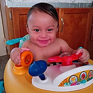 Ozias is registered to the contest to win money with this photo: baby, child, smile, happy, toy, activity_walker, indoor, kitchen, wooden_cabinet, colorful, play, person, infant, young_child, face, hands, bright, cute, joyful, home