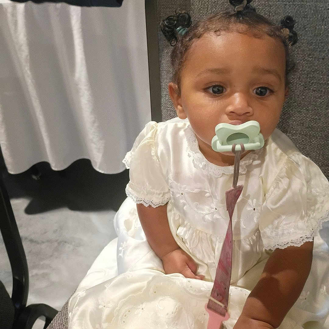 Ja'Izland is registered to the contest to win money with this photo: toddler, child, pacifier, white_dress, sitting, indoor, curly_hair, clip, face, expression, cute, person, young_child, portrait, fabric, lace, seated, baby, clothing, infant