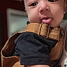 baby, infant, child, portrait, close_up, face, wide_eyes, tongue_out, smiling, indoor, warm_lighting, brown_clothing, dark_shirt, buckle, strap, adult_hand, holding, cute, selfie, family