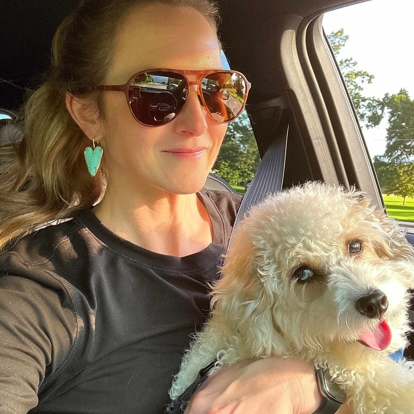 Bodie is registered to the contest to win money with this photo: woman, dog, car, sunglasses, earrings, seatbelt, sunlight, happy, pet, fluffy_dog, tongue_out, window, greenery, portrait, casual_clothing, hand, smile, interior, daylight, park