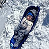Elias is registered to the contest to win money with this photo: baby, child, sled, snow, winter, snowsuit, blue, hat, outdoor, cold, snowbank, face, person, clothing, winterwear, daylight, recreation, parked_cars, buildings, play
