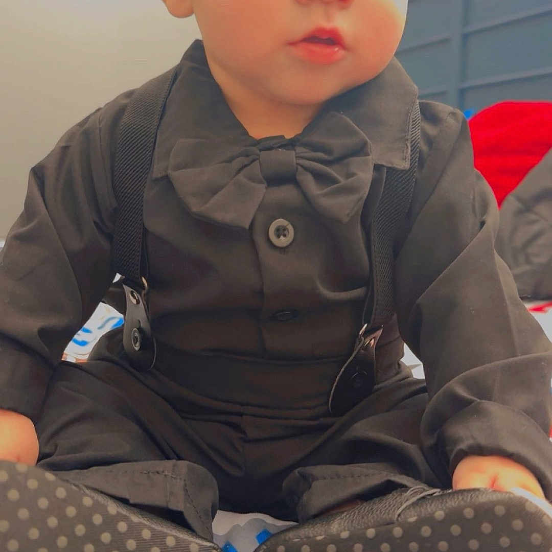 Elias is registered to the contest to win money with this photo: baby, child, infant, black_clothing, bow_tie, suspenders, sitting, indoor, blanket, footwear, sole_pattern, cute, portrait, face, eyes, young_child, person, soft_lighting, expression, baby_shoes
