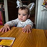 Elias is registered to the contest to win money with this photo: toddler, child, baby, animal_ears, headband, table, wood, restaurant, indoor, person, curious, clothing, long_sleeve, cute, face, hands, seat, cozy, lighting, portrait