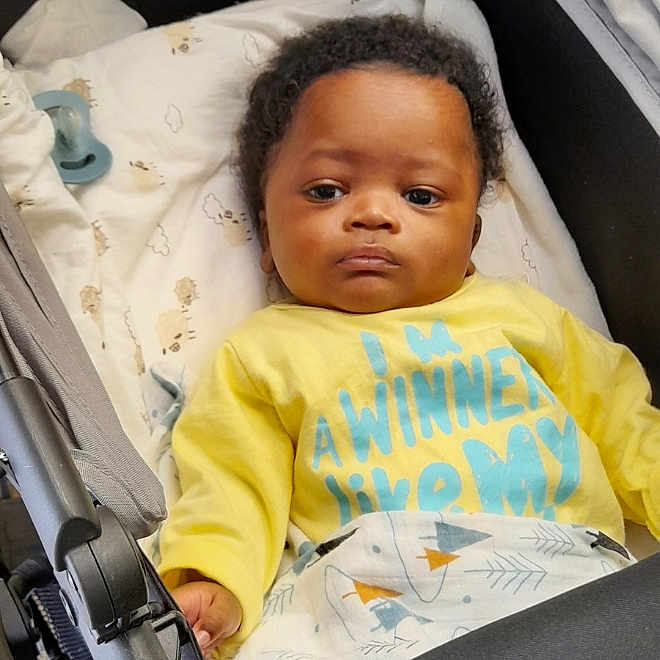 Ngom a rejoint le concours — aidez-le/la à gagner de superbes lots ! baby, baby_bed, blanket, child, clothing, comfort, curly_hair, cute, expressionless, fabric, face, indoor, infant, pacifier, person, portrait, resting, stroller, toddler, yellow_shirt