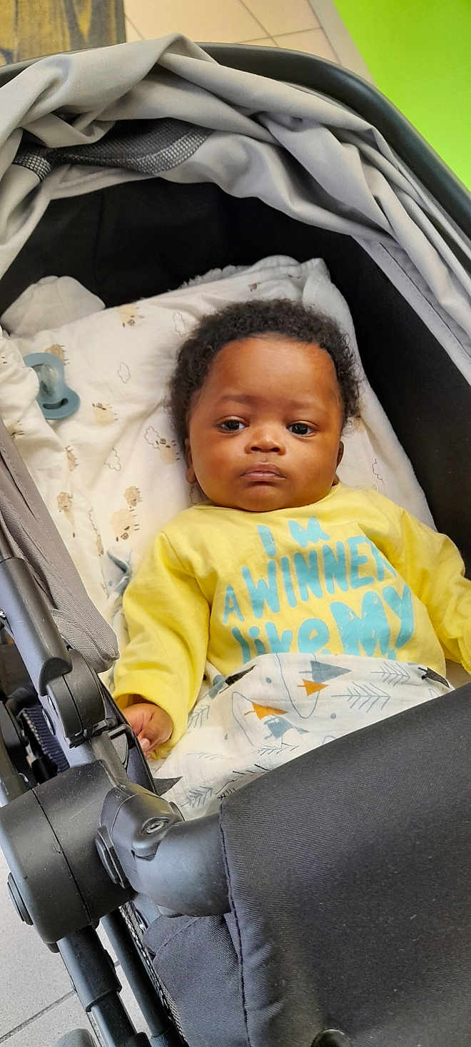 Ngom a rejoint le concours — aidez-le/la à gagner de superbes lots ! baby, infant, stroller, yellow_shirt, blanket, pacifier, face, child, indoor, expressionless, clothing, curly_hair, person, baby_bed, fabric, comfort, cute, resting, portrait, toddler