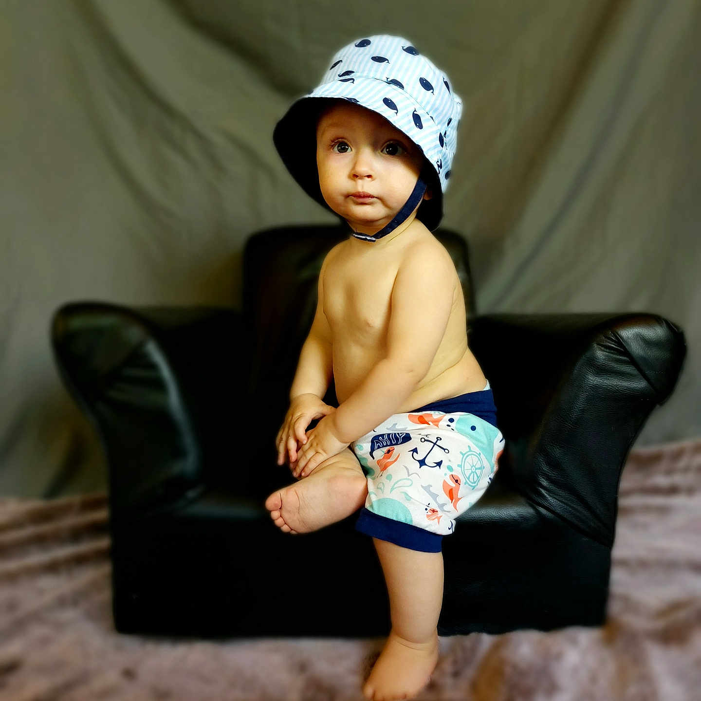 Landon is registered to the contest to win money with this photo: armchair, baby, bodypart, bonnet, cap, chair, clothing, couch, diaper, face, finger, furniture, hat, head, person, photography, portrait, shorts, sitting, sunhat