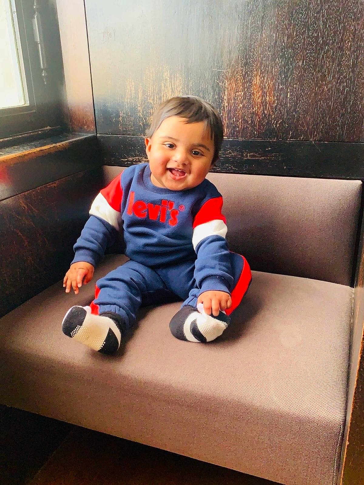 Mir is registered to the contest to win money with this photo: baby, child, smiling, sitting, bench, cushion, clothing, levis, window, wood, indoor, happy, person, face, footwear, sock, cute, portrait, cozy, natural_light
