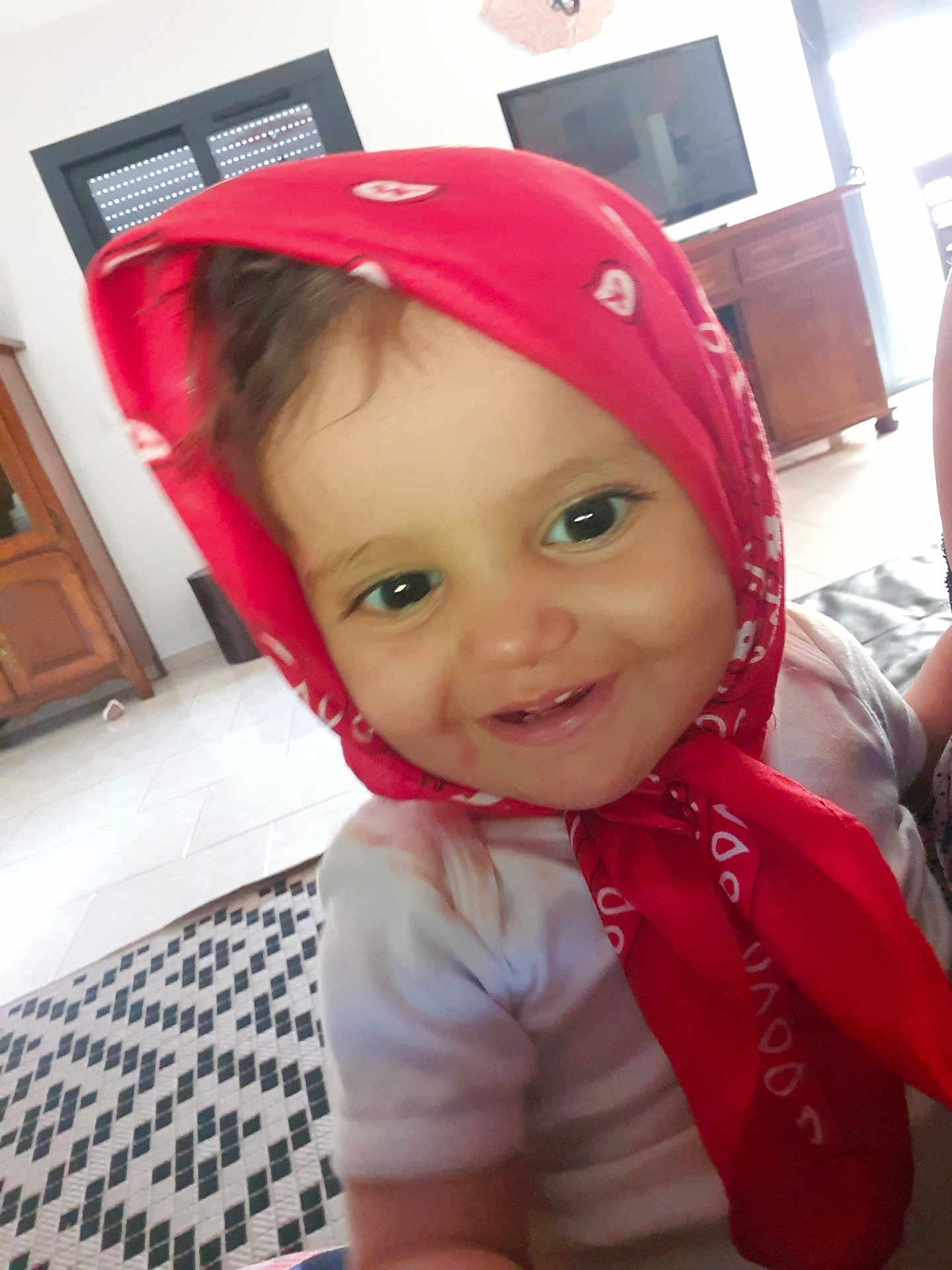 Salma a rejoint le concours — aidez-le/la à gagner de superbes lots ! baby, child, toddler, red_bandana, smile, face, indoor, furniture, television, carpet, wooden_cabinet, shirt, cheerful, young_child, headwear, person, home, closeup, cute, portrait