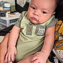 baby, infant, child, person, onesie, green_clothing, hand, adult_hand, face, portrait, indoor, cozy_room, furniture, casual_clothing, cute, chubby_cheeks, sitting, holding, skin, closeup