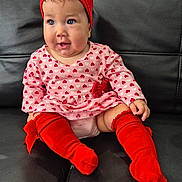 Aisha Hechvarria joined the competition — help win amazing prizes! baby, child, infant, red_headband, red_socks, pink_dress, hearts_pattern, sitting, black_couch, cute, happy, portrait, indoor, head_accessory, footwear, person, small_child, toddler, smile, fashion