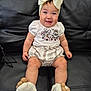 baby, child, smiling, sitting, bow, headwear, blue_eyes, white_shirt, patterned_shirt, striped_shorts, socks, flower_socks, leather_couch, indoor, cute, happy, portrait, infant, person, human