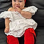 baby, child, red_bow, headband, white_dress, red_socks, couch, leather, sitting, cute, playful, finger_near_mouth, portrait, indoors, fashion, smiling, happy, infant, clothing, person