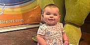 Avalyn is registered to the contest to win money with this photo: baby, child, smiling, floor, dress, floral_pattern, mascot, costume, legs, tile, indoor, colorful, background, person, happy, seated, cute, infant, playful, advertisement