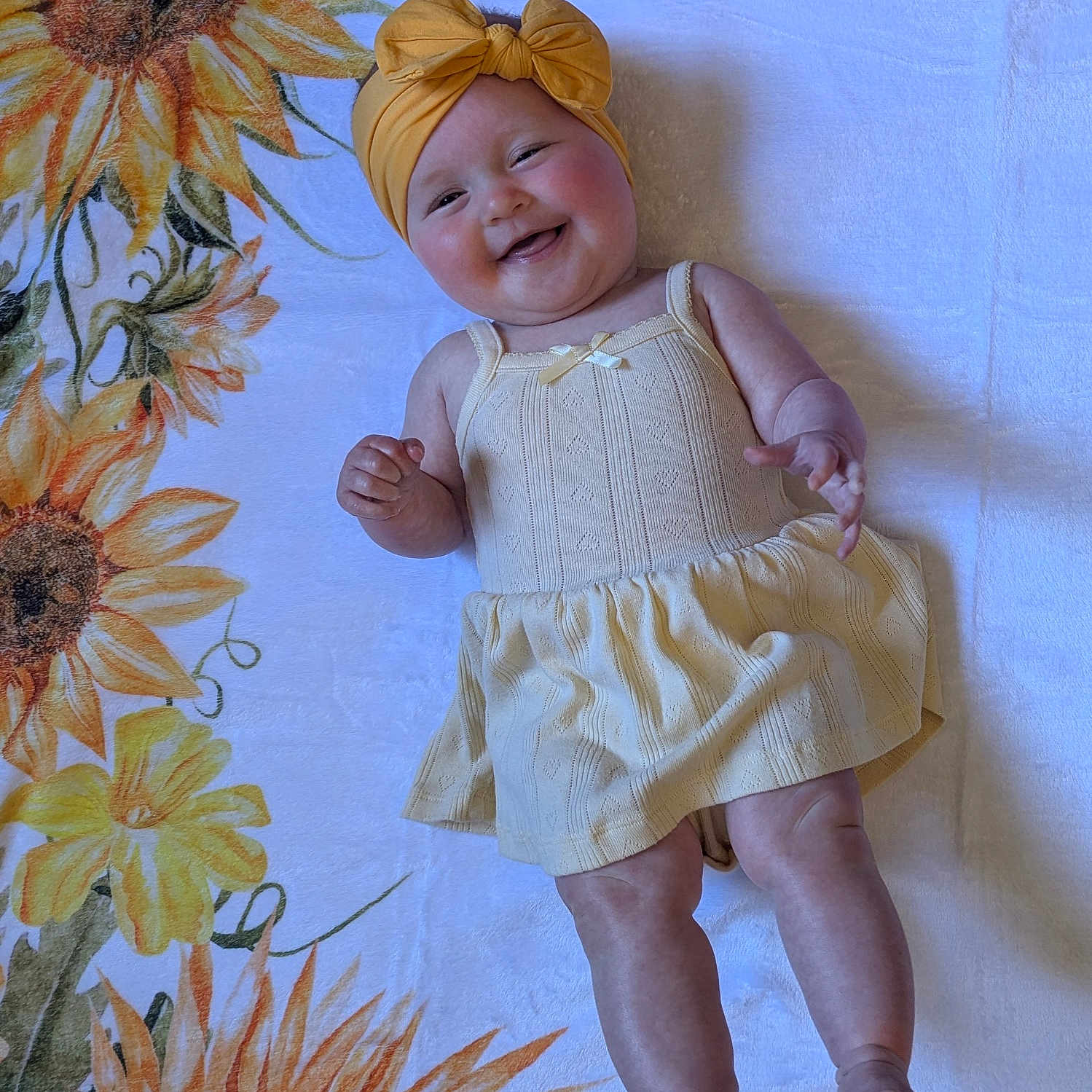 Eloise joined the competition — help win amazing prizes! arms, baby, blanket, bow, child, cute, dress, face, happy, headband, indoors, infant, legs, lying_down, pattern, skin, smiling, soft_texture, sunflower, yellow