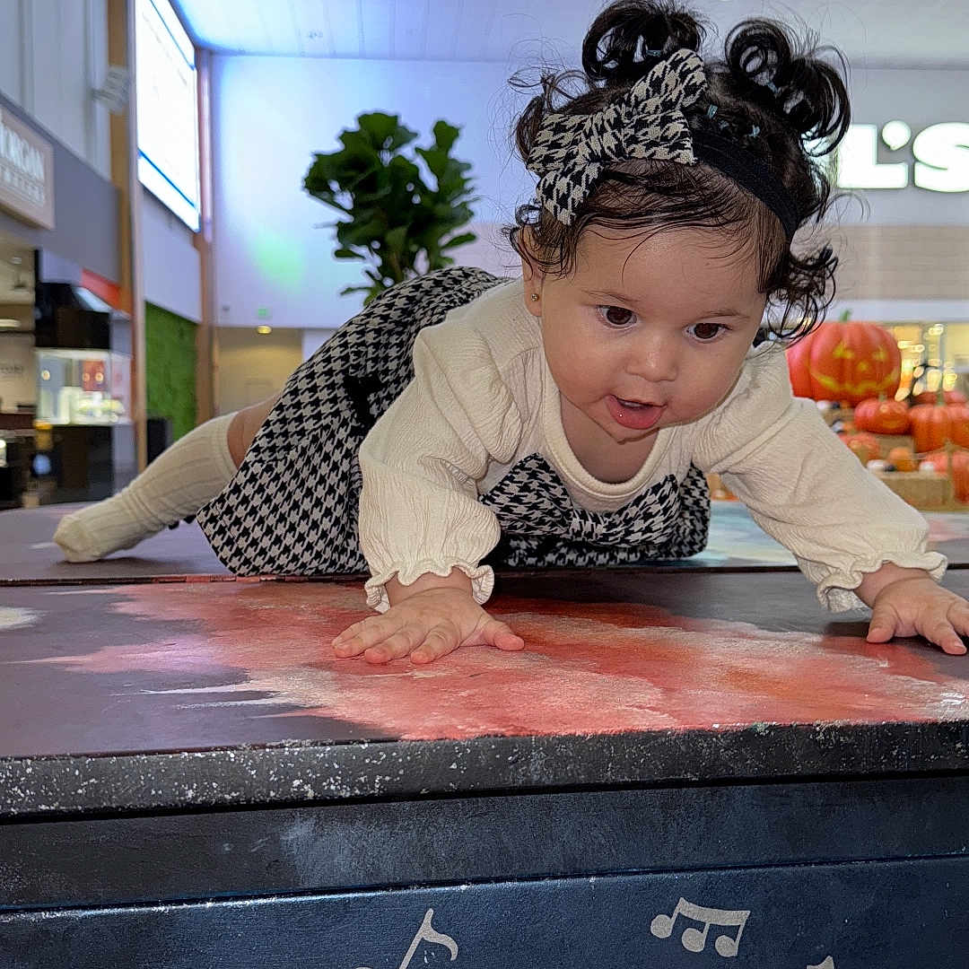 Julieth Camila Novoa joined the competition — help win amazing prizes! bow, child, crawling, curiosity, curly_hair, face, floor, hands, headband, indoor, lighting, long_sleeve, mall, music_notes, patterned_clothing, person, plant, pumpkin, table, toddler