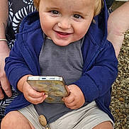 Jagger is registered to the contest to win money with this photo: toddler, child, smiling, phone, blue_hoodie, shorts, sitting, outdoor, bench, gravel, person, hands, face, hair, playful, cute, happy, casual_clothing, wood, toy_chain