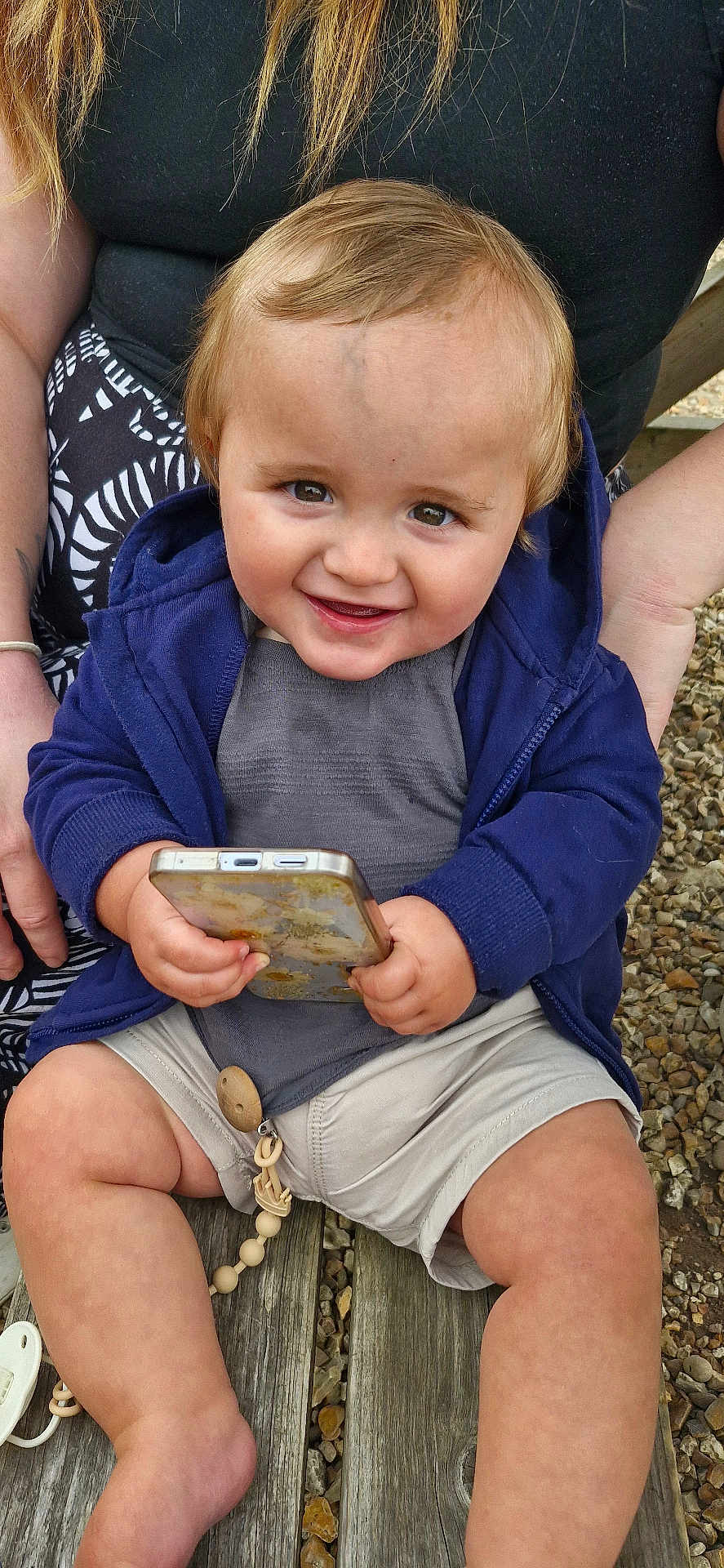 Jagger is registered to the contest to win money with this photo: toddler, child, smiling, phone, blue_hoodie, shorts, sitting, outdoor, bench, gravel, person, hands, face, hair, playful, cute, happy, casual_clothing, wood, toy_chain