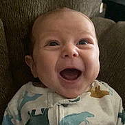 Levi is registered to the contest to win money with this photo: baby, infant, smiling, happy, onesie, dinosaur_print, clothing, face, head, sitting, couch, indoor, child, cute, portrait, person, expression, young, comfort, cozy