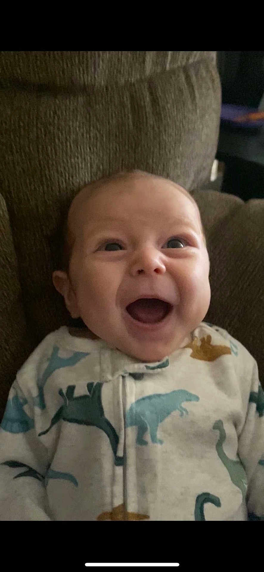Levi is registered to the contest to win money with this photo: baby, infant, smiling, happy, onesie, dinosaur_print, clothing, face, head, sitting, couch, indoor, child, cute, portrait, person, expression, young, comfort, cozy