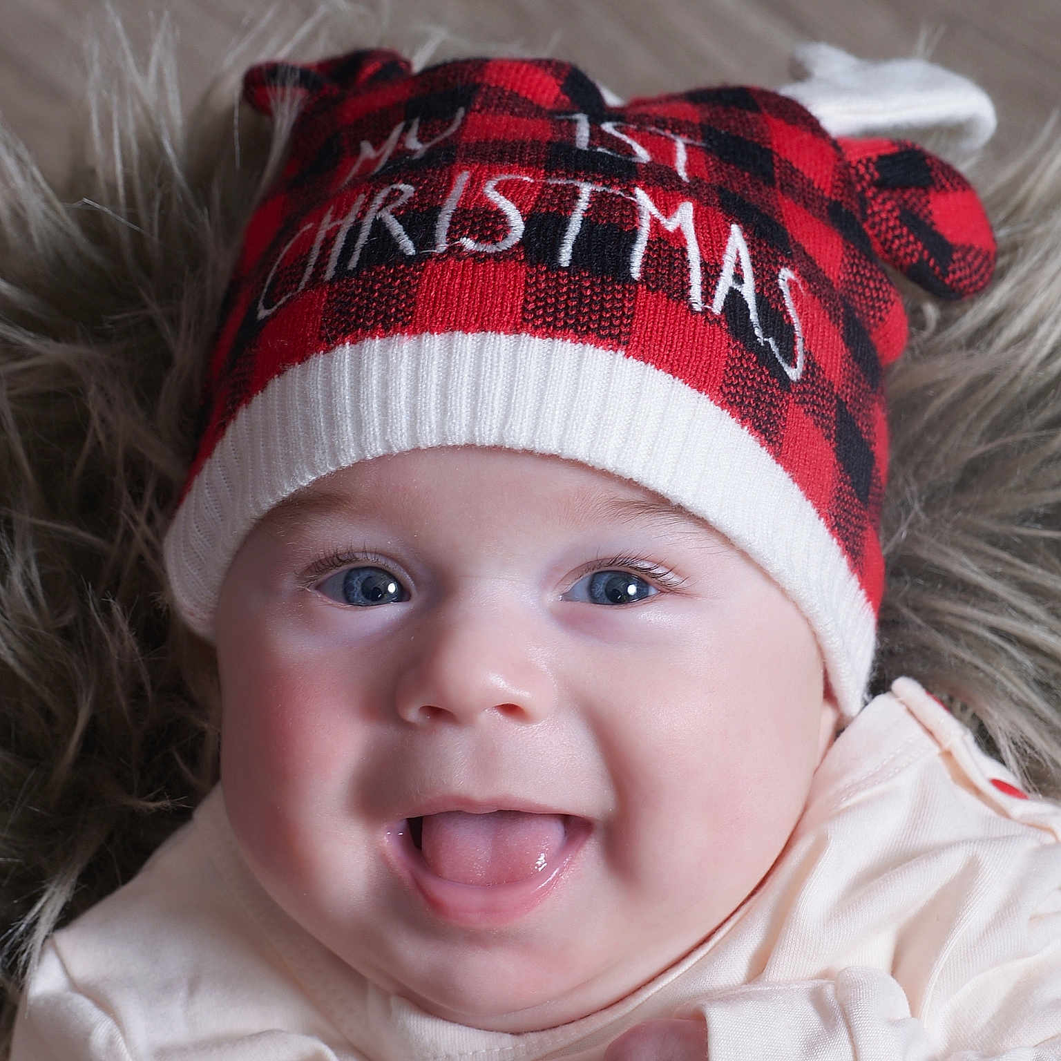 Leonidas is registered to the contest to win money with this photo: baby, black, blue_eyes, cheeks, child, closeup, clothing, cute, face, fur_background, happy, hat, infant, plaid, portrait, red, smiling, soft, tongue_out, warm