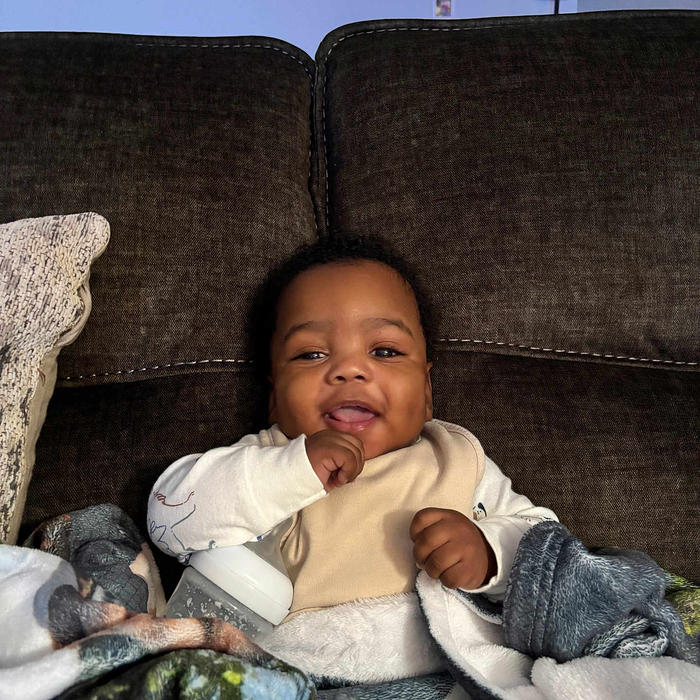 Karay is registered to the contest to win money with this photo: baby, infant, smiling, bottle, blanket, couch, indoor, cozy, child, portrait, happy, wall_art, photoframes, home, furniture, lamp, casual_clothing, young_child, people, relaxing