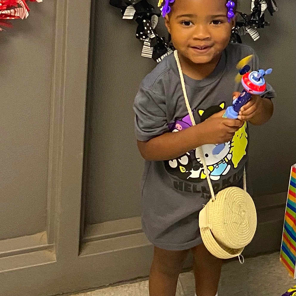 Corina is registered to the contest to win money with this photo: basket_purse, black_hair, child, colorful, cute, decor, floor_tiles, gray_tshirt, hair_bows, hello_kitty, indoor, person, playful, ribbon_garland, smiling, sneakers, standing, toddler, toy, white_socks