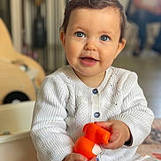 Alice participe au concours pour gagner de l'argent avec cette photo : baby, child, smiling, blue_eyes, white_sweater, toy, orange_toy, indoor, seated, cute, happy, portrait, infant, knitted_clothing, hands, patterned_pants, home, blurred_background, floor, person