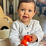 baby, child, smiling, blue_eyes, white_sweater, toy, orange_toy, indoor, seated, cute, happy, portrait, infant, knitted_clothing, hands, patterned_pants, home, blurred_background, floor, person