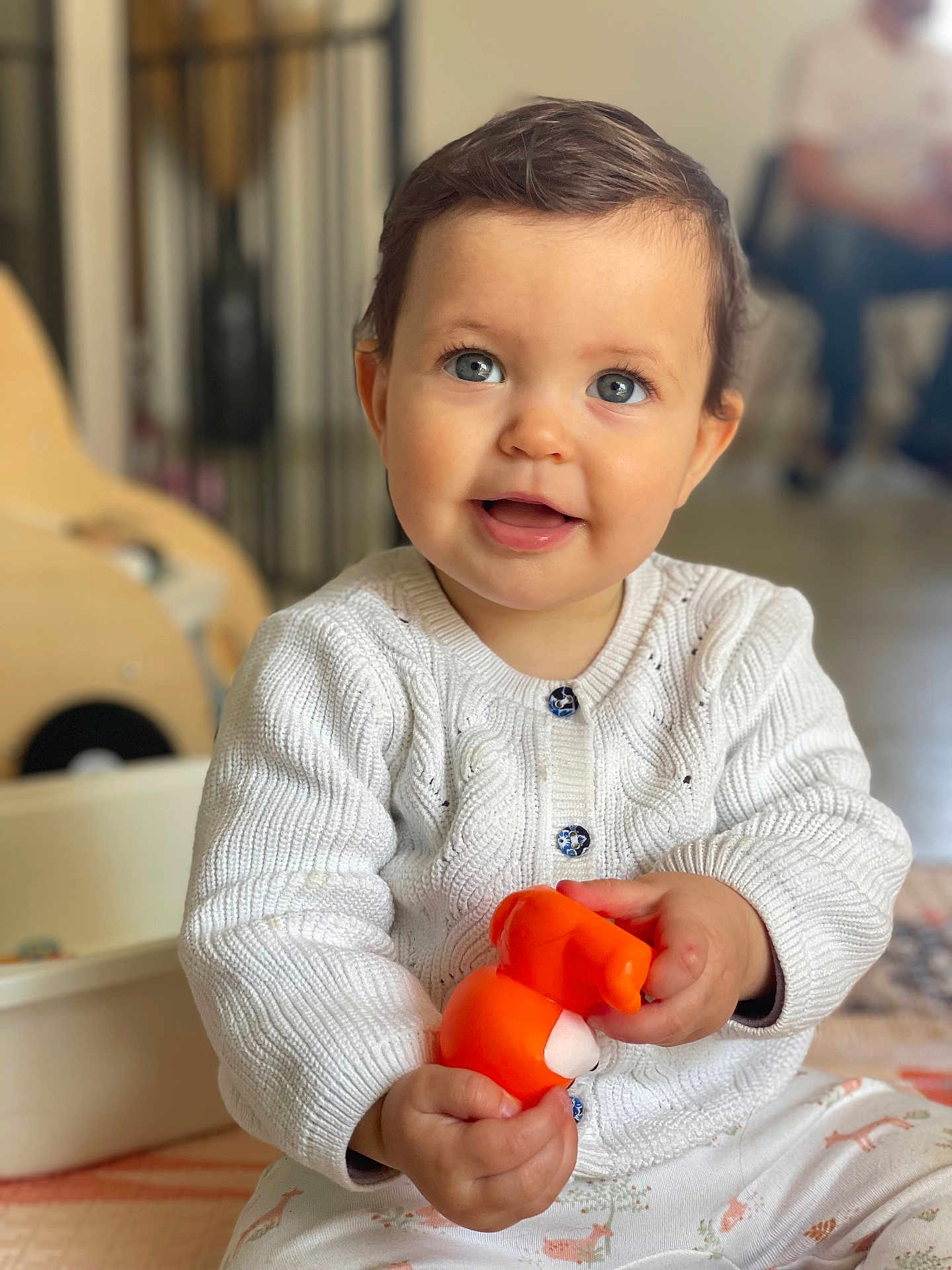 Alice participe au concours pour gagner de l'argent avec cette photo : baby, child, smiling, blue_eyes, white_sweater, toy, orange_toy, indoor, seated, cute, happy, portrait, infant, knitted_clothing, hands, patterned_pants, home, blurred_background, floor, person