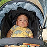 baby, child, stroller, outdoor, portrait, sitting, serious, face, brown_hair, dinosaur_shirt, yellow_clothing, gray_pants, infant, canopy, baby_carriage, harness, toy, teether, sunlight, photography