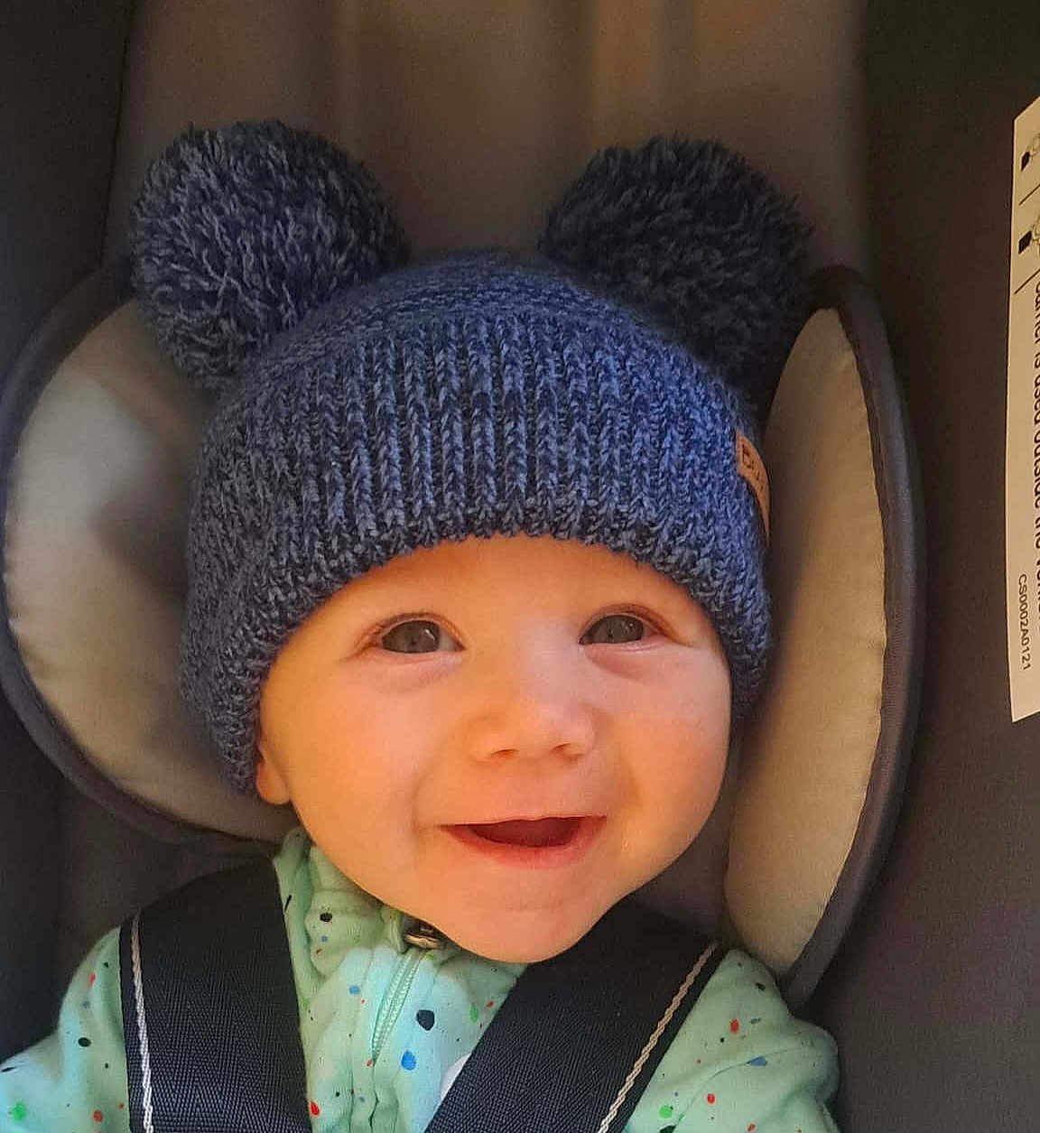 Noah is registered to the contest to win money with this photo: baby, child, smiling, hat, knit_hat, pom_poms, car_seat, seatbelt, cute, happy, infant, face, clothing, portrait, indoors, young_child, headwear, apparel, person, closeup