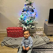Serhat a rejoint le concours — aidez-le/la à gagner de superbes lots ! blanket, child, christmas_tree, clothing, curious, decorations, floor, furniture, gift_box, holiday, home, indoor, lights, ornaments, person, present, rug, sitting, smiling, toddler