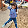 child, girl, smiling, posing, blue_top, jeans, sneakers, outdoor, playground, wood_chips, sidewalk, trees, people, park, daylight, casual_clothing, happy, fun, nature, recreation