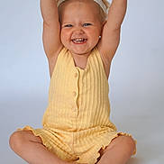 Lina a rejoint le concours — aidez-le/la à gagner de superbes lots ! toddler, child, smiling, yellow_clothing, headband, barefoot, sitting, raising_arms, happy, studio, portrait, cute, baby, indoor, light_background, short_hair, buttoned_outfit, skin, feet, hands