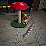 cat, orange_tabby, scratching_post, mushroom_shaped, feather_toy, colorful_toy, fuzzy_toy, indoor, tile_floor, chair, cardboard_box, curious, playful, pet_toy, animal, feline, crouching, home, cozy, yellow_string