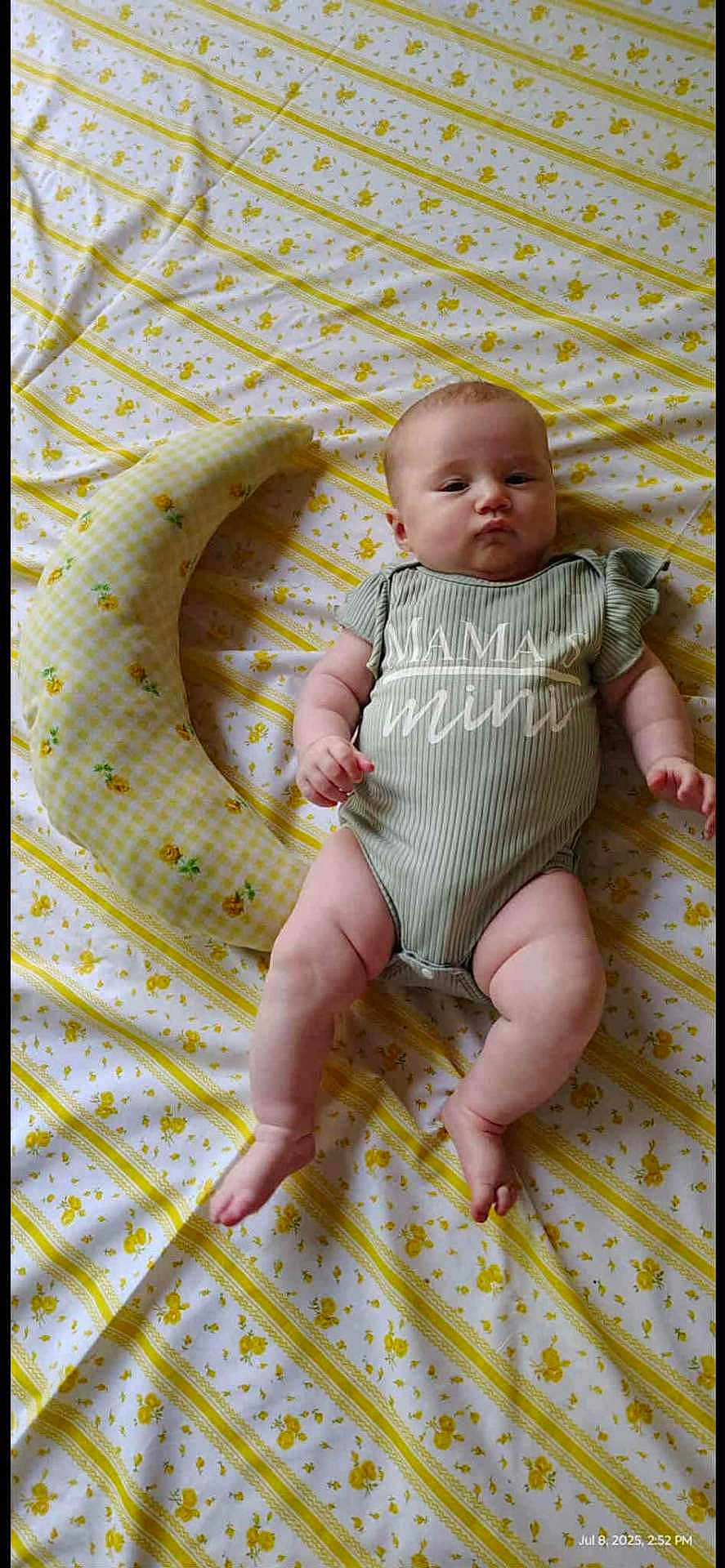 Kinsleigh is registered to the contest to win money with this photo: baby, infant, onesie, green_clothing, yellow_flower_pattern, crescent_pillow, bed_sheet, lying_down, face, cute, child, indoors, soft_toy, person, newborn, pillow, patterned_fabric, skin, expression, resting