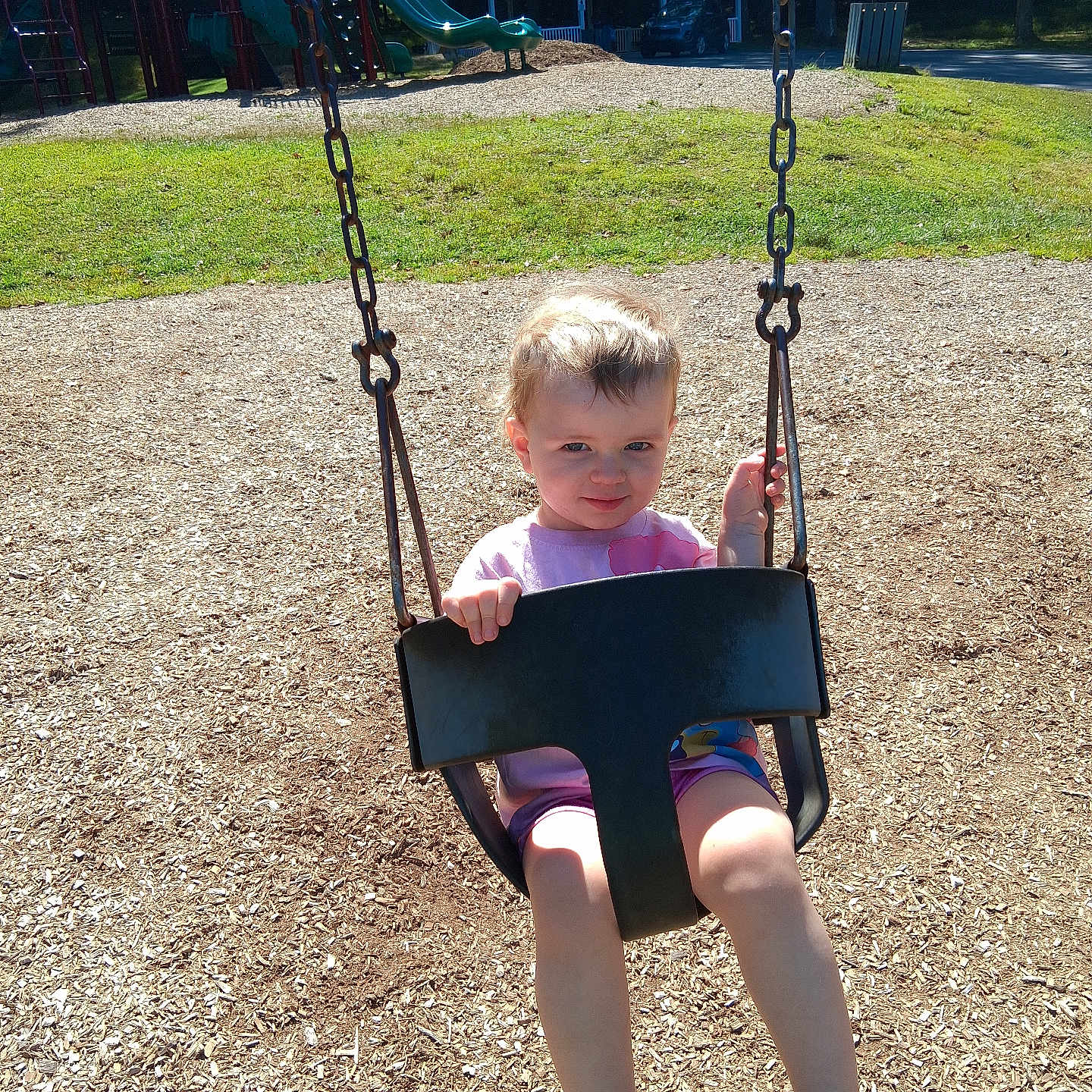 Bryleigh is registered to the contest to win money with this photo: car, casual_clothing, child, daylight, footwear, grass, outdoor, park, play_equipment, playground, safety, shirt, shorts, smile, summer, sunlight, swing, toddler, trees, woodchips