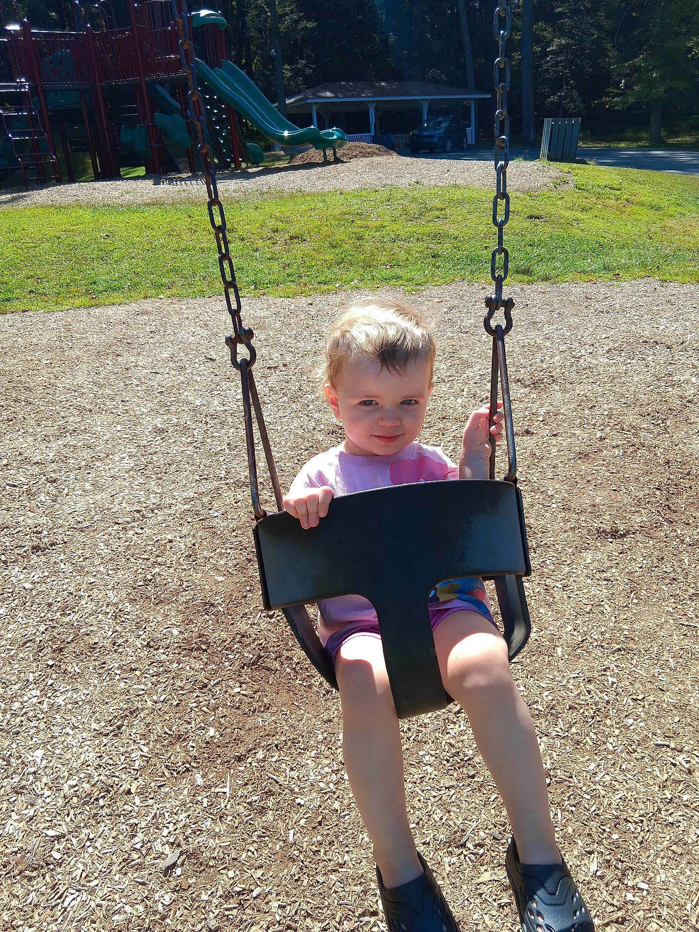 Bryleigh is registered to the contest to win money with this photo: child, toddler, swing, playground, outdoor, daylight, smile, woodchips, play_equipment, park, summer, casual_clothing, shorts, shirt, footwear, sunlight, grass, trees, car, safety