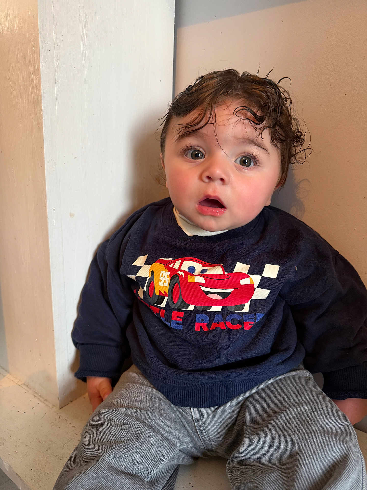 Noah participe au concours pour gagner de l'argent avec cette photo : toddler, child, sitting, curly_hair, wet_hair, surprised_expression, sweatshirt, race_car_graphic, navy_blue, gray_pants, indoor, corner, wall, casual_clothing, young_child, portrait, cute, big_eyes, small_hands, light_skin