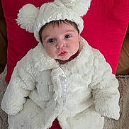 Harper is registered to the contest to win money with this photo: baby, infant, child, white_coat, pom_pom_hat, cute, portrait, indoor, red_pillow, fuzzy_clothing, warm_clothing, seated, looking_at_camera, face, eyes, cozy, soft_texture, adorable, young_child, headwear