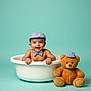 Krishika joined the competition — help win amazing prizes! baby, teddy_bear, tub, cap, bow_tie, smiling, child, toy, cute, portrait, happy, indoor, blue_background, plush, sitting, clothing, infant, accessory, studio, playful