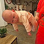 arm, baby, clothing, drying_rack, expression, face, floor, furniture, garland, hand, holding, holiday_decor, home, indoor, infant, person, plant, red_shirt, table, tattoo