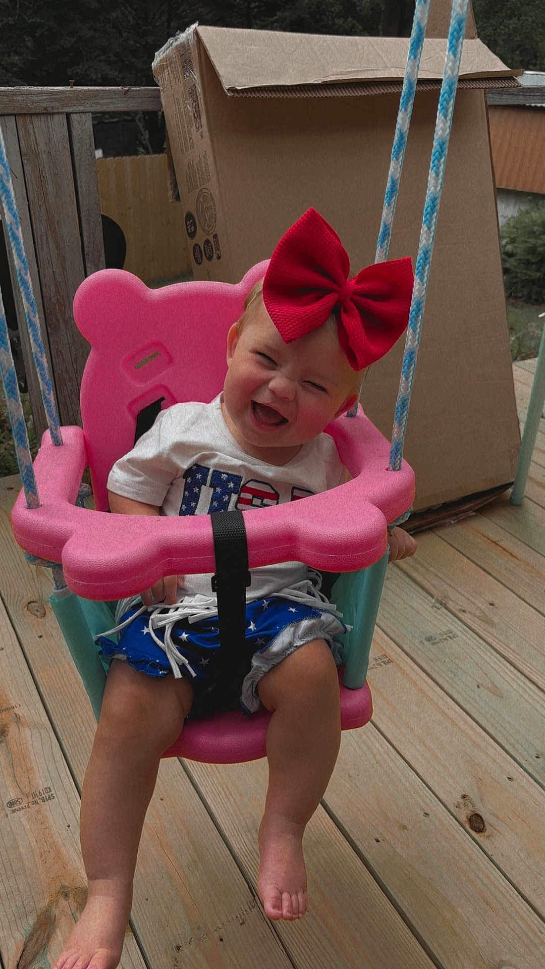Paityn is registered to the contest to win money with this photo: toddler, child, swing, pink_swing, red_bow, smiling, happy, outdoor, wooden_deck, cardboard_box, summer_clothes, barefoot, playtime, fun, rope, seat, person, baby, cute, cheerful