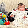 baby, infant, onesie, plush_toy, elephant_toy, patchwork_dog, pillow, blanket, smiling, cute, indoors, soft_texture, patterned, child, comfort, playful, happy, seated, portrait, cozy