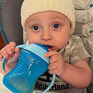 Braxton is registered to the contest to win money with this photo: baby, blue_sippy_cup, child, closeup, clothing, curious, cute, expression, face, feeding, hands, high_chair, indoor, infant, knit_hat, person, plastic_cup, portrait, seated, table
