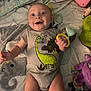 baby, blanket, bright, child, clothing, colorful, cute, dinosaur_onesie, face, hand_toy, happy, indoors, infant, laying_down, person, play_mat, plush_toy, smiling, soft, toy