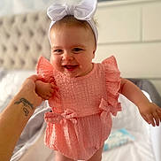 Josie-Leigh joined the competition — help win amazing prizes! baby, child, smiling, pink_dress, headband, white_bow, holding_hand, bedroom, blurred_background, happy, infant, cute, standing, person, indoor, skin, face, apparel, tattoo, adult_hand