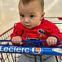 baby, child, shopping_cart, red_sweater, batman_logo, gray_pants, indoor, hands, face, eyes, infant, clothing, person, portrait, grip, cute, curious, young_child, retail, store
